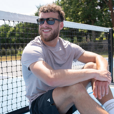 Lifestyle image of a model sitting on a pickleball court and wearing a pair of JOOLA RJX Lite eyewear.