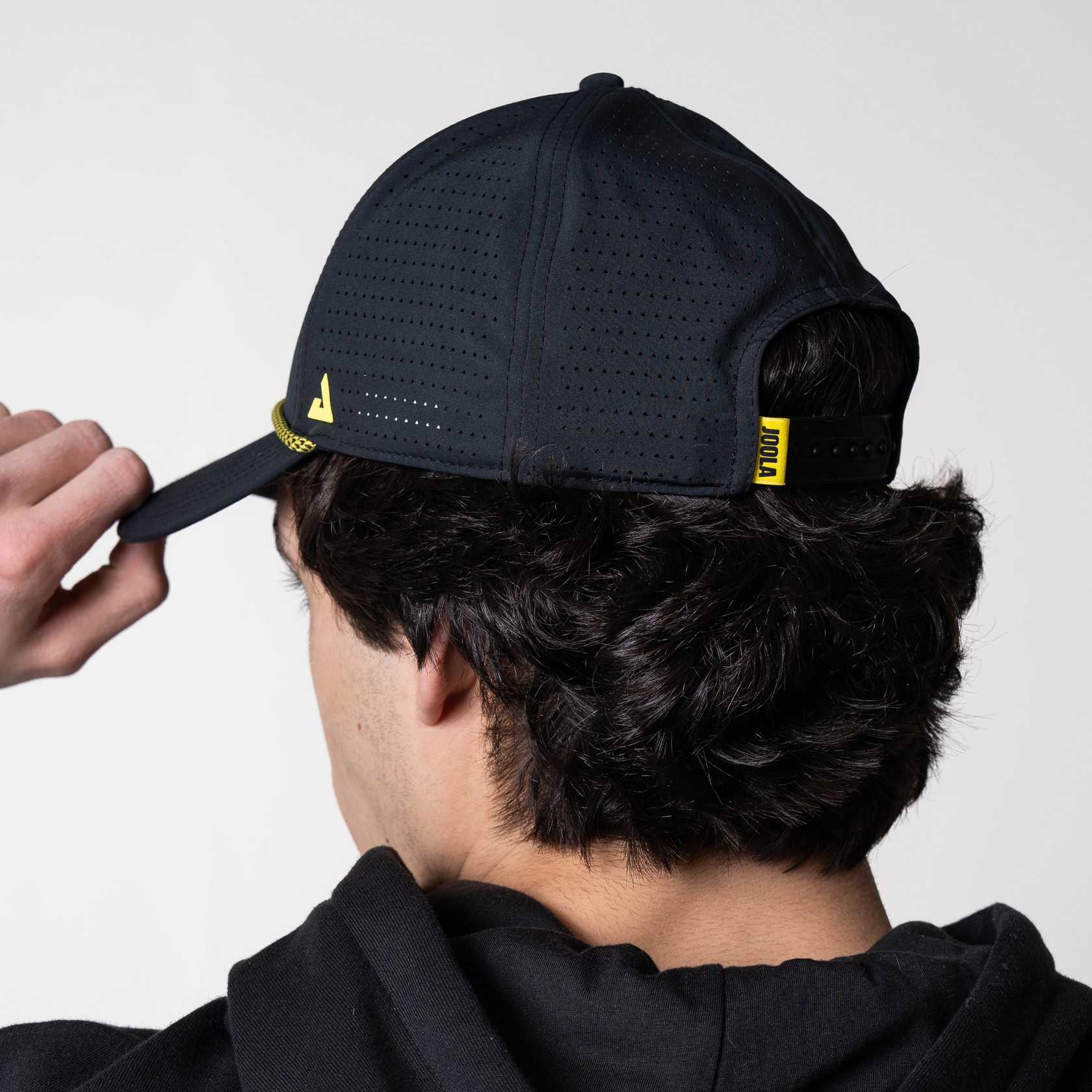 White background, image of the back of a JOOLA Rope Cap, black with yellow branded logos.