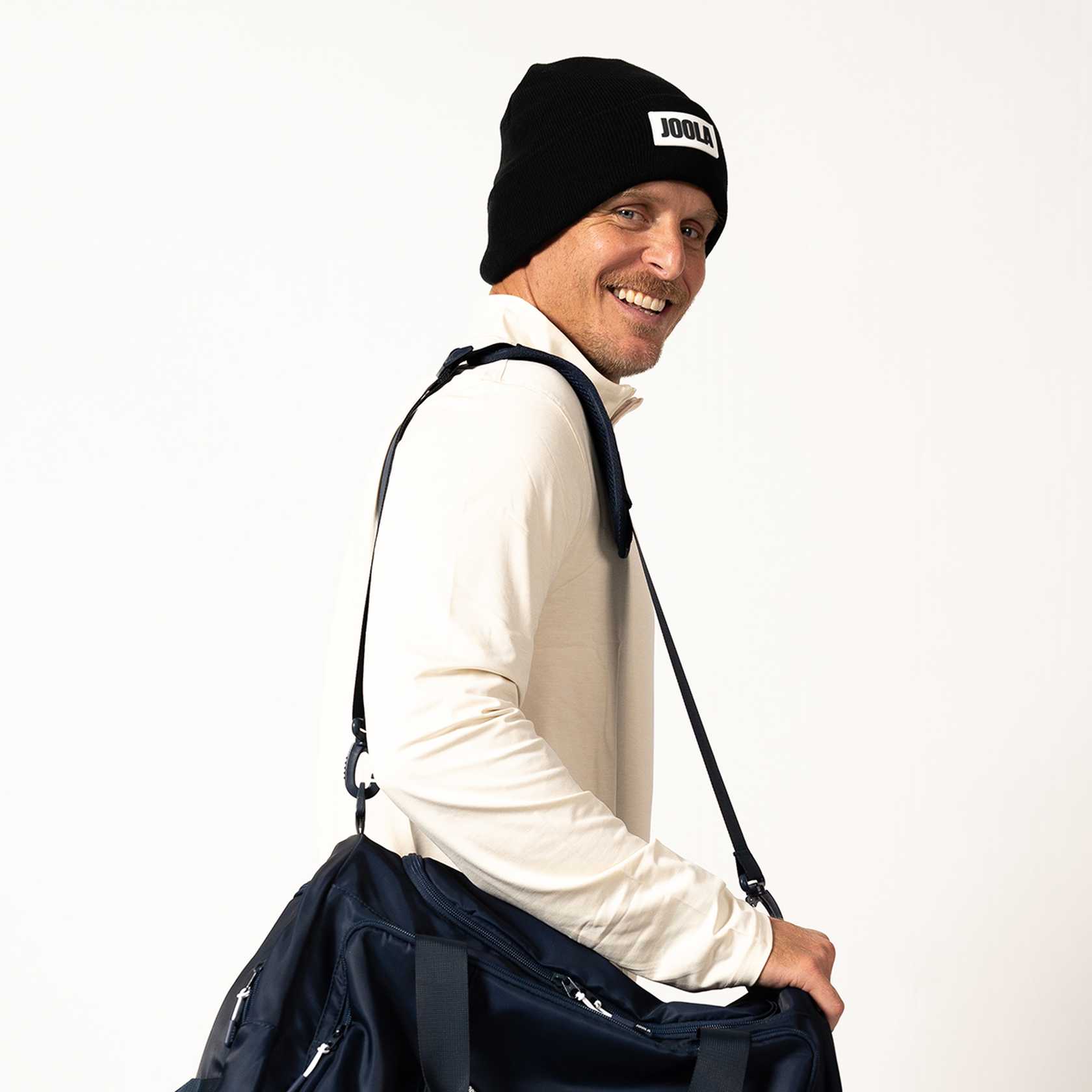 Man wearing a black beanie with 'JOOLA' logo and holding a navy bag on a white background