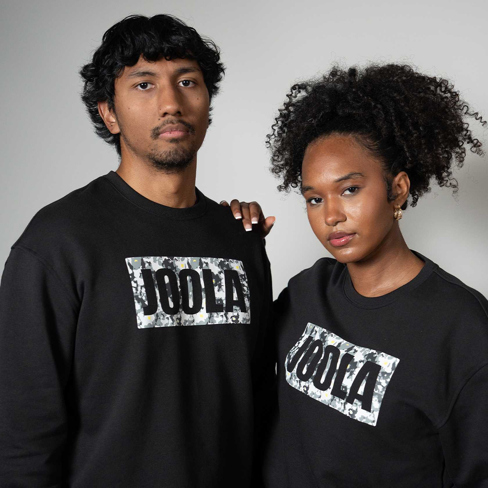 Two people wearing black fleece JOOLA sweatshirts with a brand logo on a gray background
