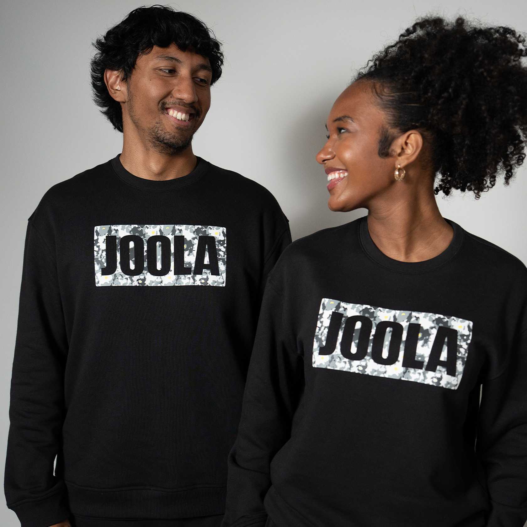 Two people wearing black fleece sweatshirts with 'JOOLA' logo on a gray background.