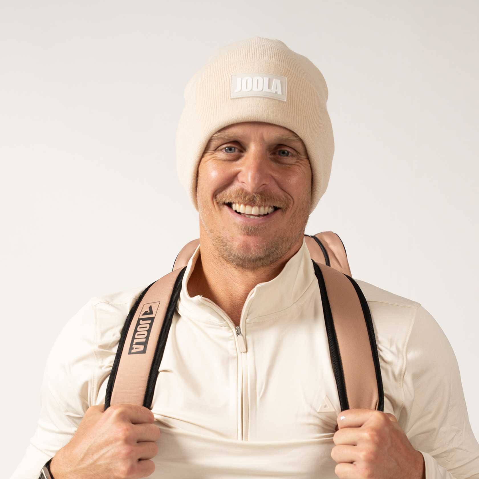 Man wearing a JOOLA Latte beanie and holding a pink backpack with JOOLA branding on a white background