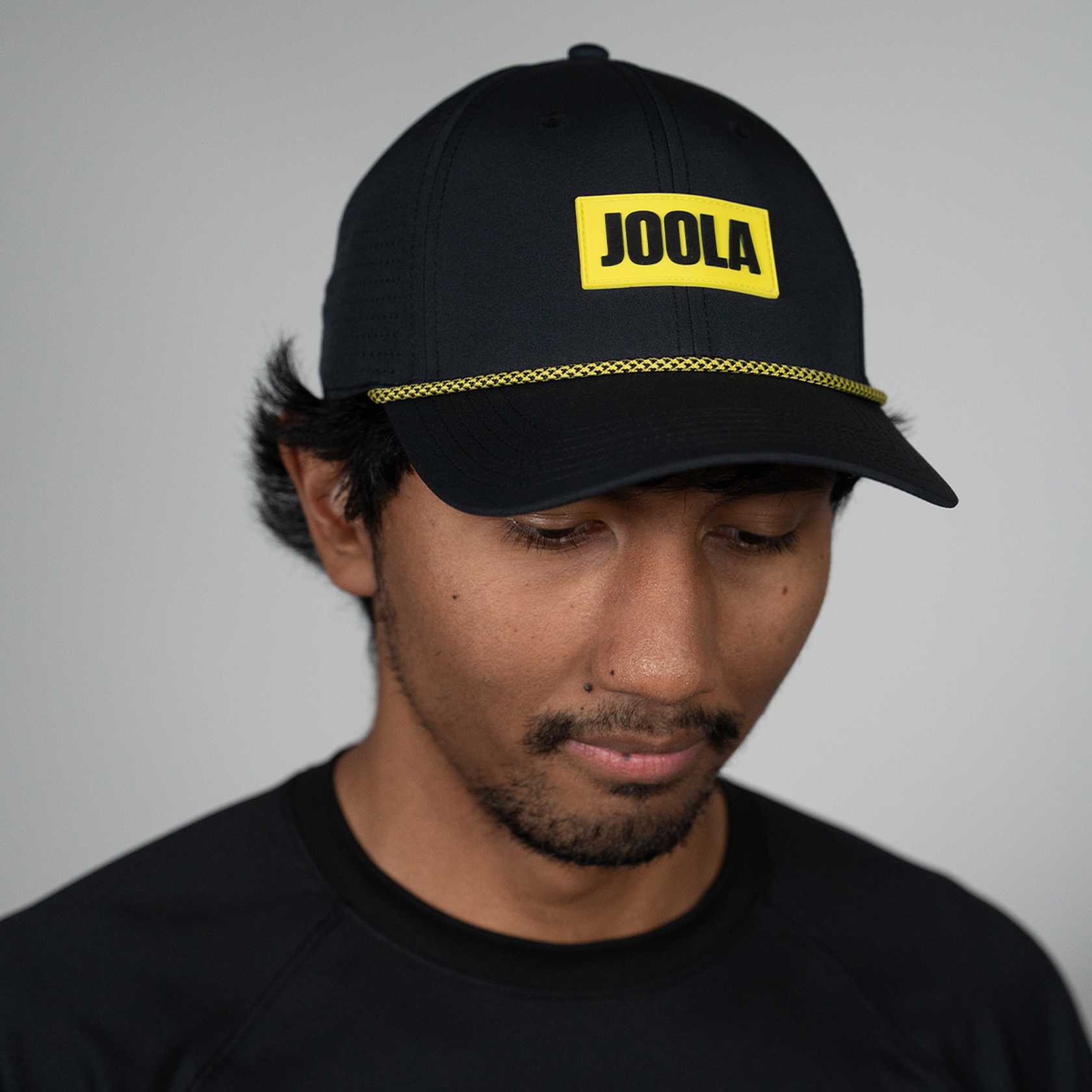 Person wearing a black cap with 'JOOLA' logo on a gray background, black and yellow hat.