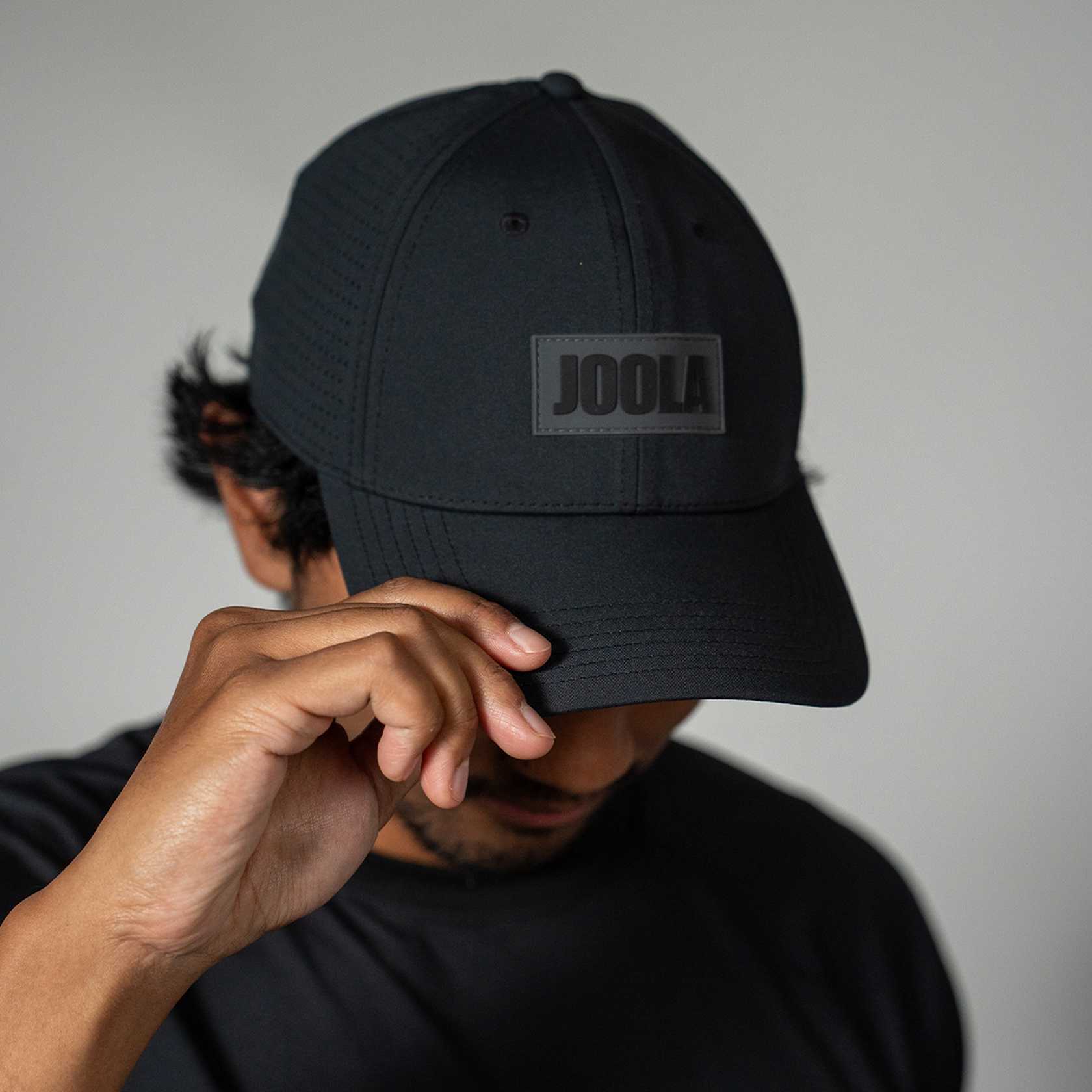 Person wearing a black cap with 'JOOLA' logo against a neutral background