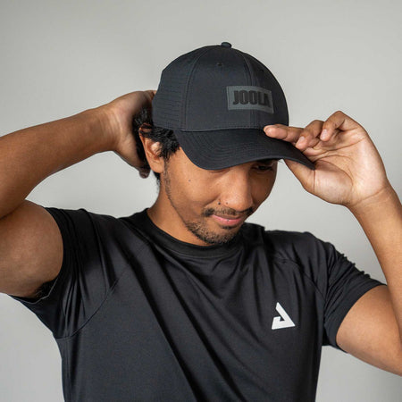 Man wearing a black cap with 'JOOLA' logo and black t-shirt with white logo on gray background