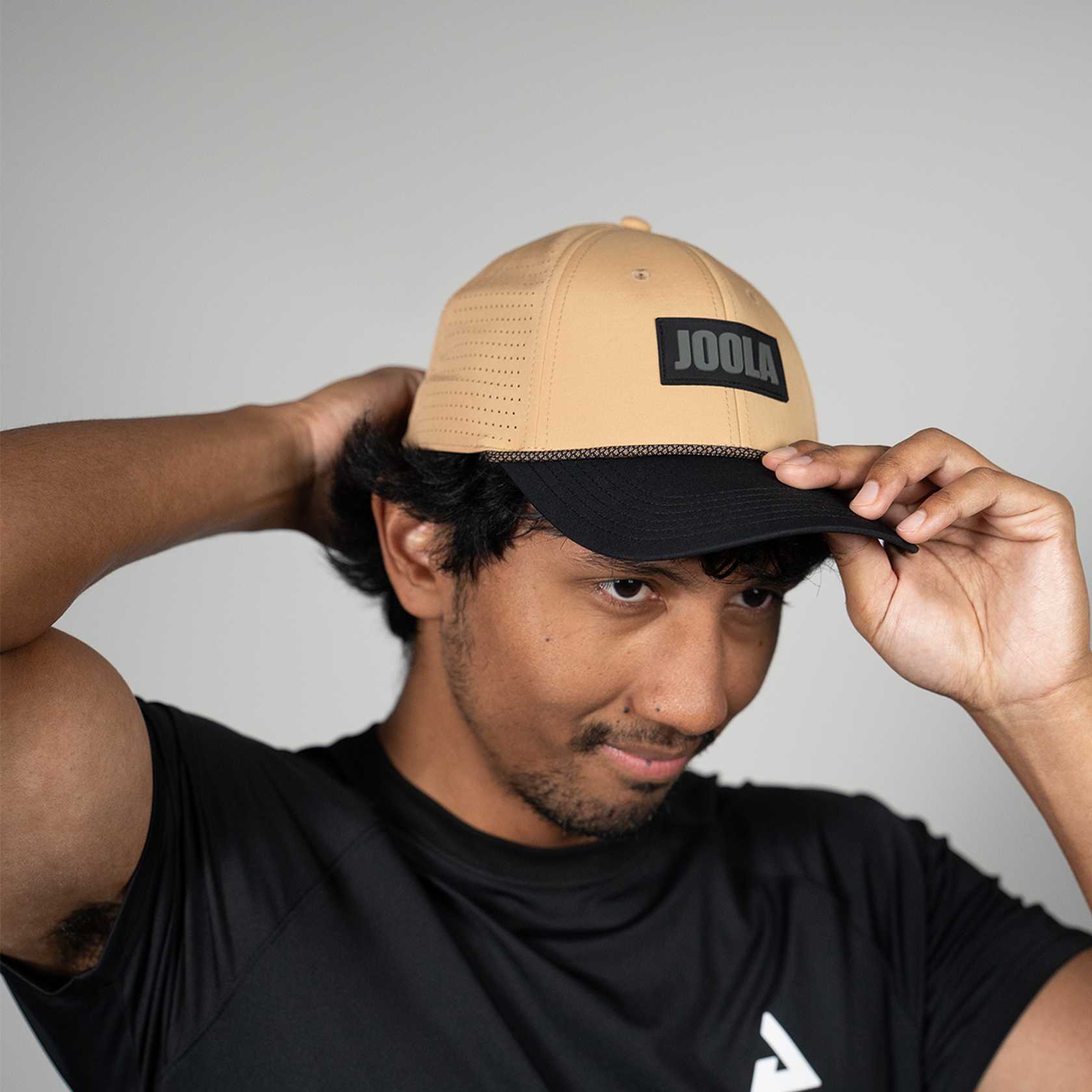 Person wearing a latte and black cap with 'JOOLA' branding against a gray background.