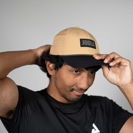 Person wearing a latte and black cap with 'JOOLA' branding against a gray background.