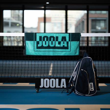 Image of a green JOOLA wordmark towel hanging over a JOOLA pickleball net.