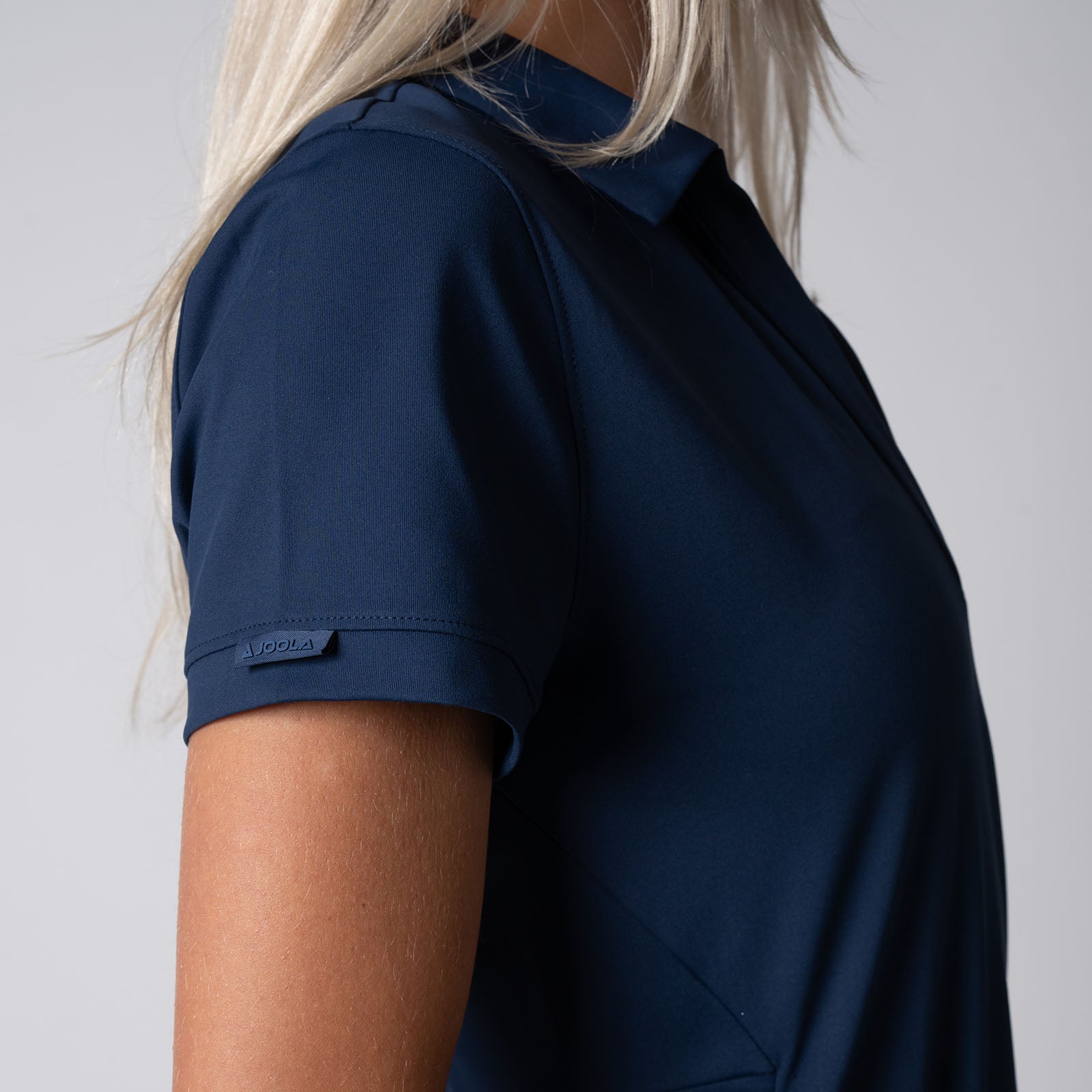 Close up image showing the JOOLA logo tag on the right sleeve of a JOOLA Women's Club Polo Dress, Navy.