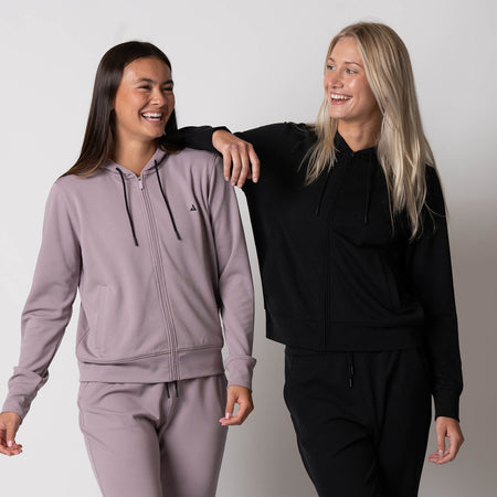 Two female models wearing JOOLA women's ribbed jackets. The women's on the left is wearing a dusty mauve colored jacket and the women on the right is wearing a black JOOLA Women's Ribbed jacket.