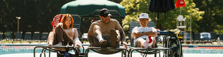 Three people relax in lounge chairs by a swimming pool outdoors, with trees, umbrellas, and poolside equipment in the background.