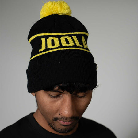 Person wearing a black beanie with yellow JOOLA text and pom-pom on a gray background