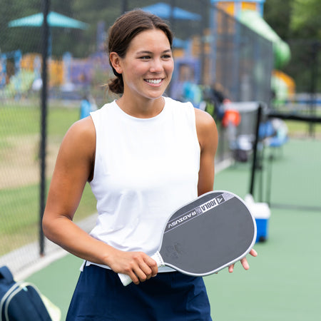 A woman smiling on a pickleball court, wearing a white sleeveless top and a navy blue skirt, holding a paddle. A navy backpack is on the court beside her.