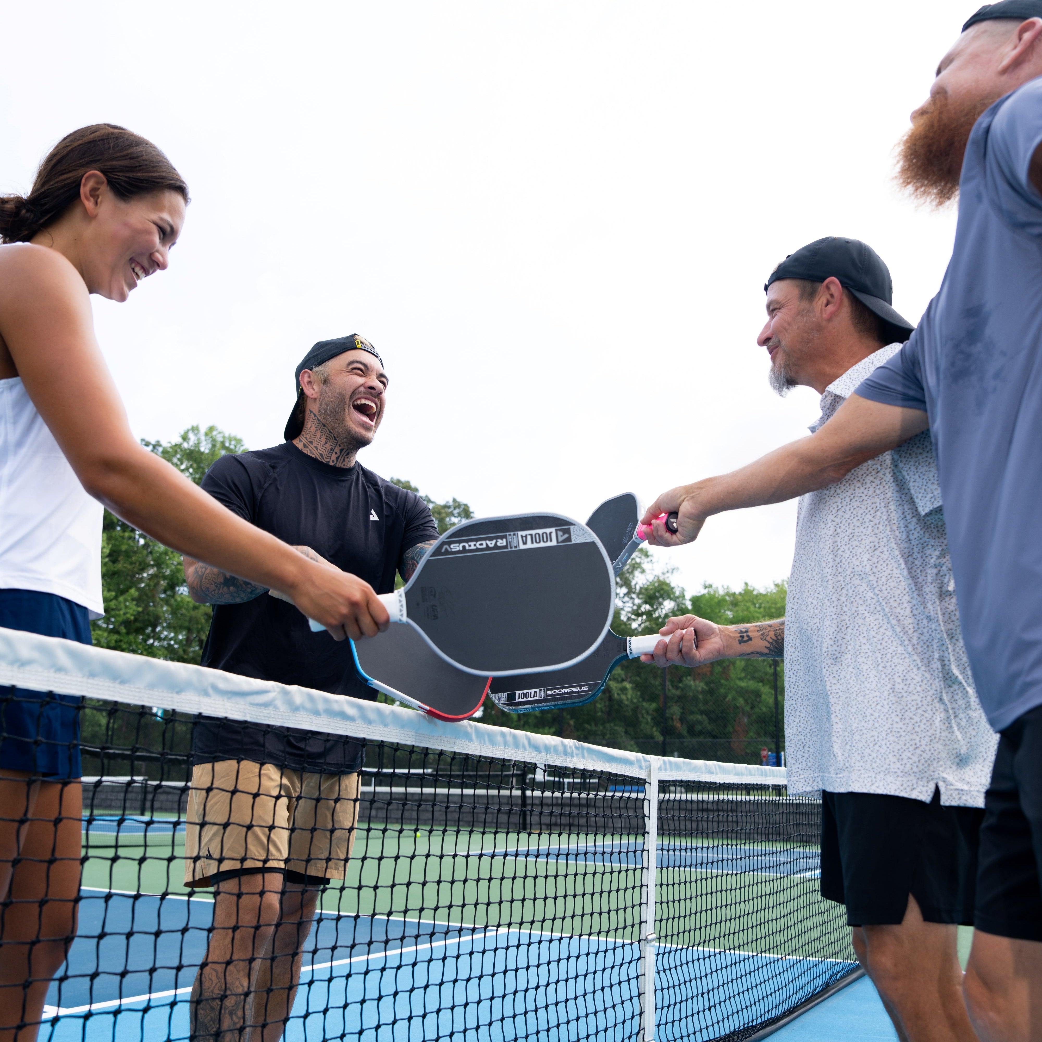 Four people on a blue pickleball court shaking hands over a net, with one person holding a black and gray paddle. The scene is outdoors on a tennis/pickleball court with trees in the background.