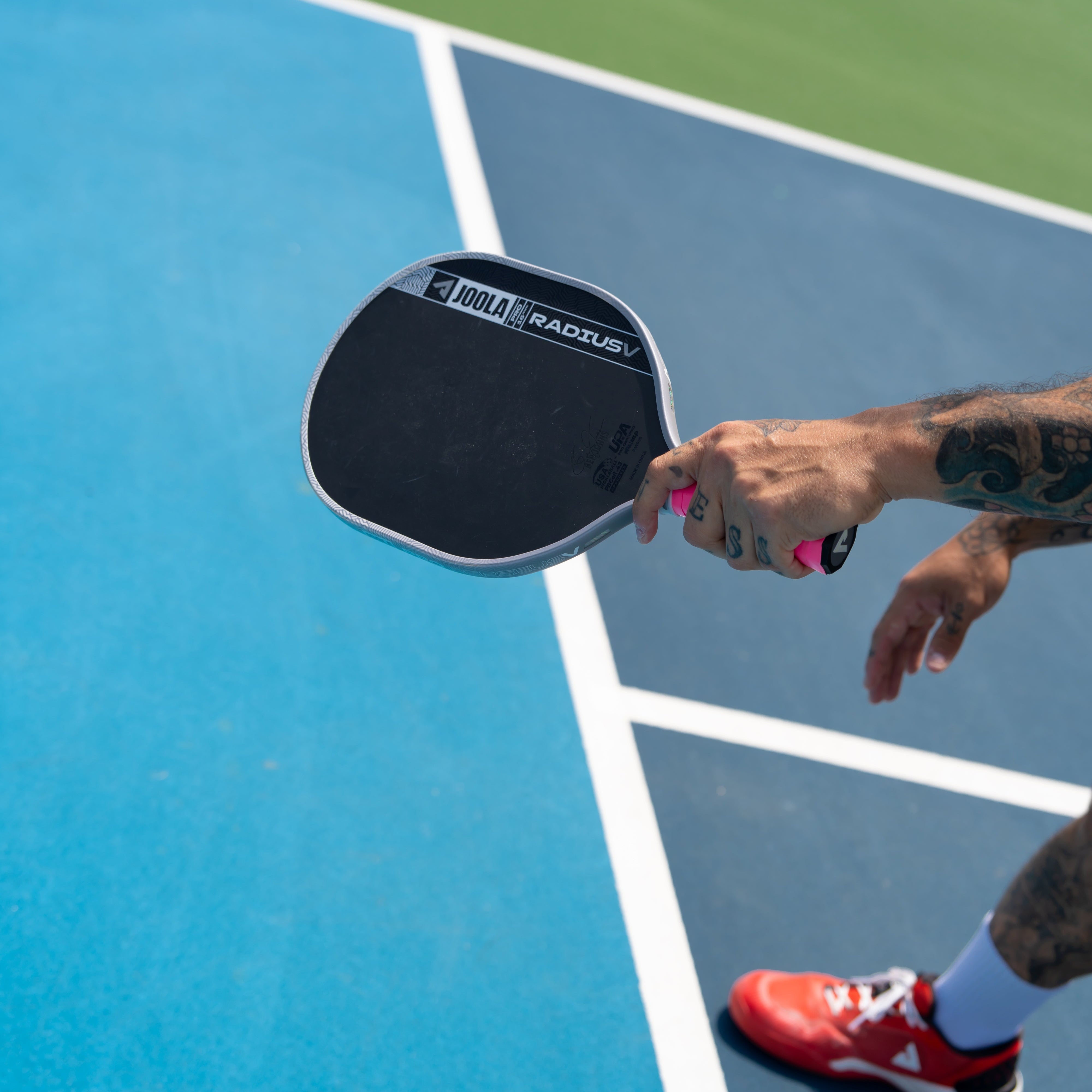 A close-up action shot of a person holding a pickleball paddle on a blue and green pickleball court. The paddle has a black face with a white and black 'Pickleball' logo.